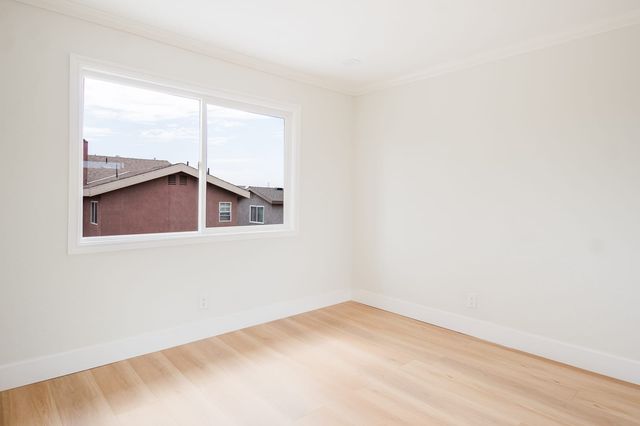 An empty bedroom with light wood plank flooring, white baseboards and a sliding window looking out at neighboring stucco rooftops