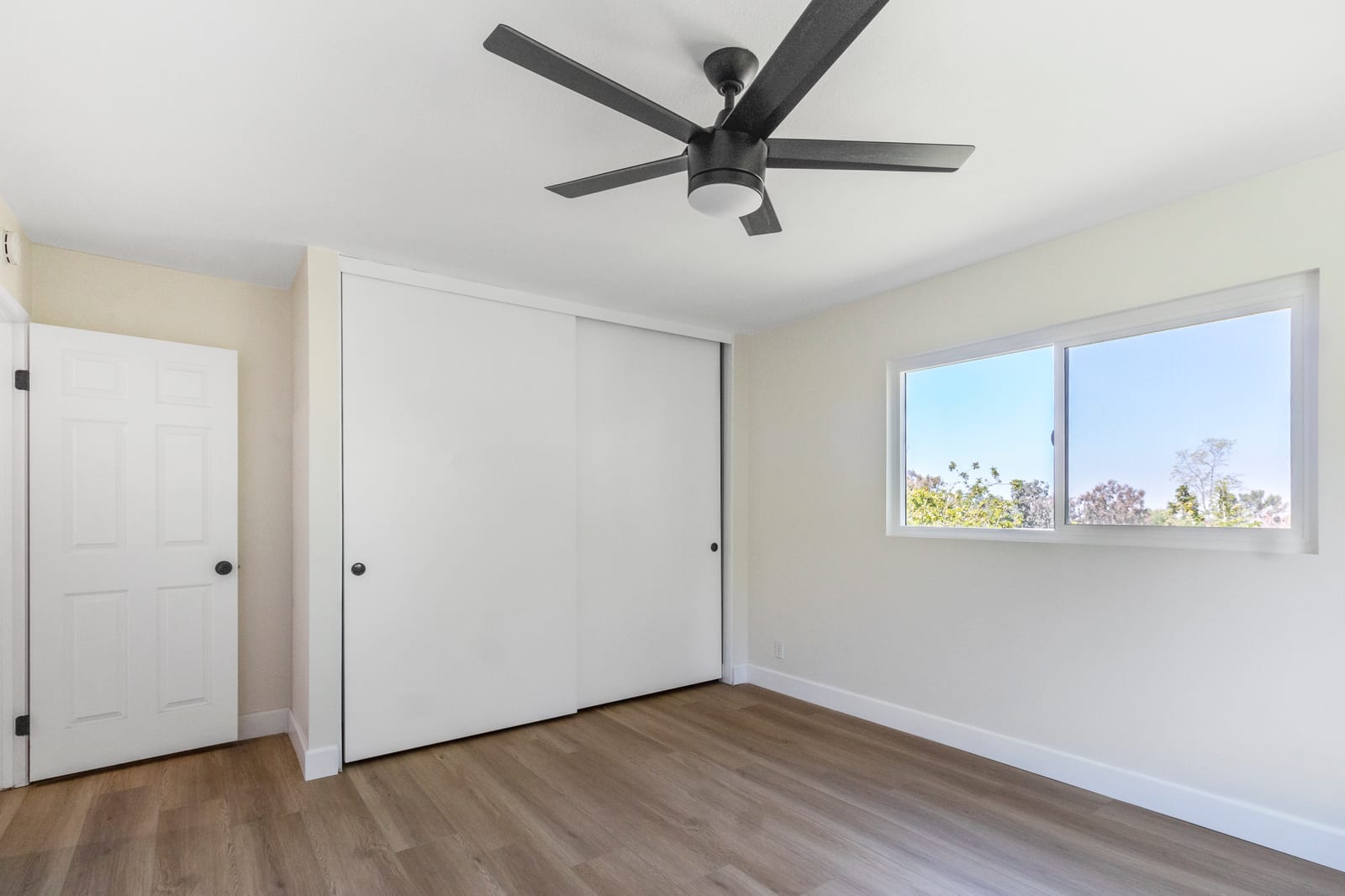 Empty bedroom with wood-look plank flooring, white sliding closet doors, a black ceiling fan and a window looking out to trees