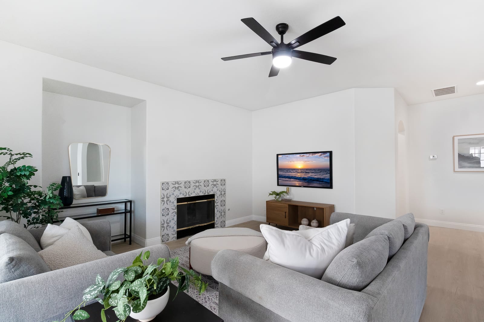 Staged living room with gray sofas, a patterned tile fireplace surround, a wall-mounted television and a black ceiling fan