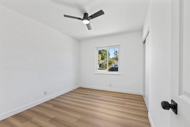 Empty bedroom with light wood plank flooring, a black ceiling fan and a window looking out to a residential street