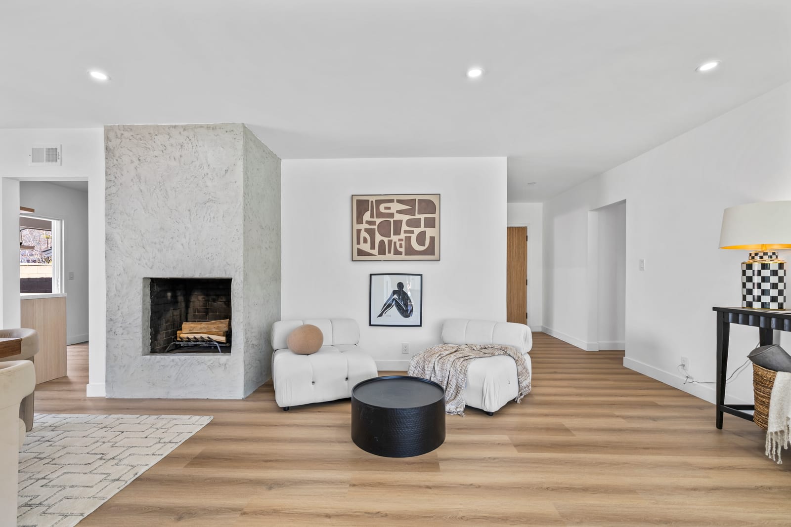 Living room with a textured plaster fireplace, two white lounge chairs, a round black table and wood plank floors