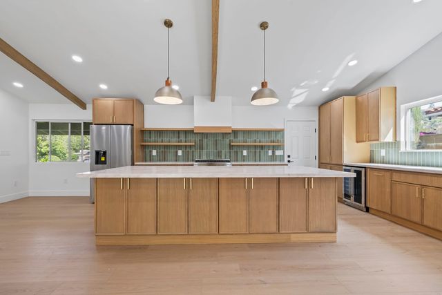 Kitchen in a remodeled Claremont home: a long wood-cabinet island with a pale stone top under two hanging pendant lights, exposed ceiling beams, and a green tile backsplash with open shelves