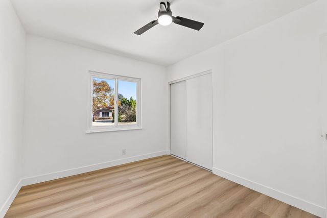 Empty bedroom in a Claremont home with light wood-look flooring, a white sliding closet door, a ceiling fan, and a window looking out on trees and a single-story house across the street