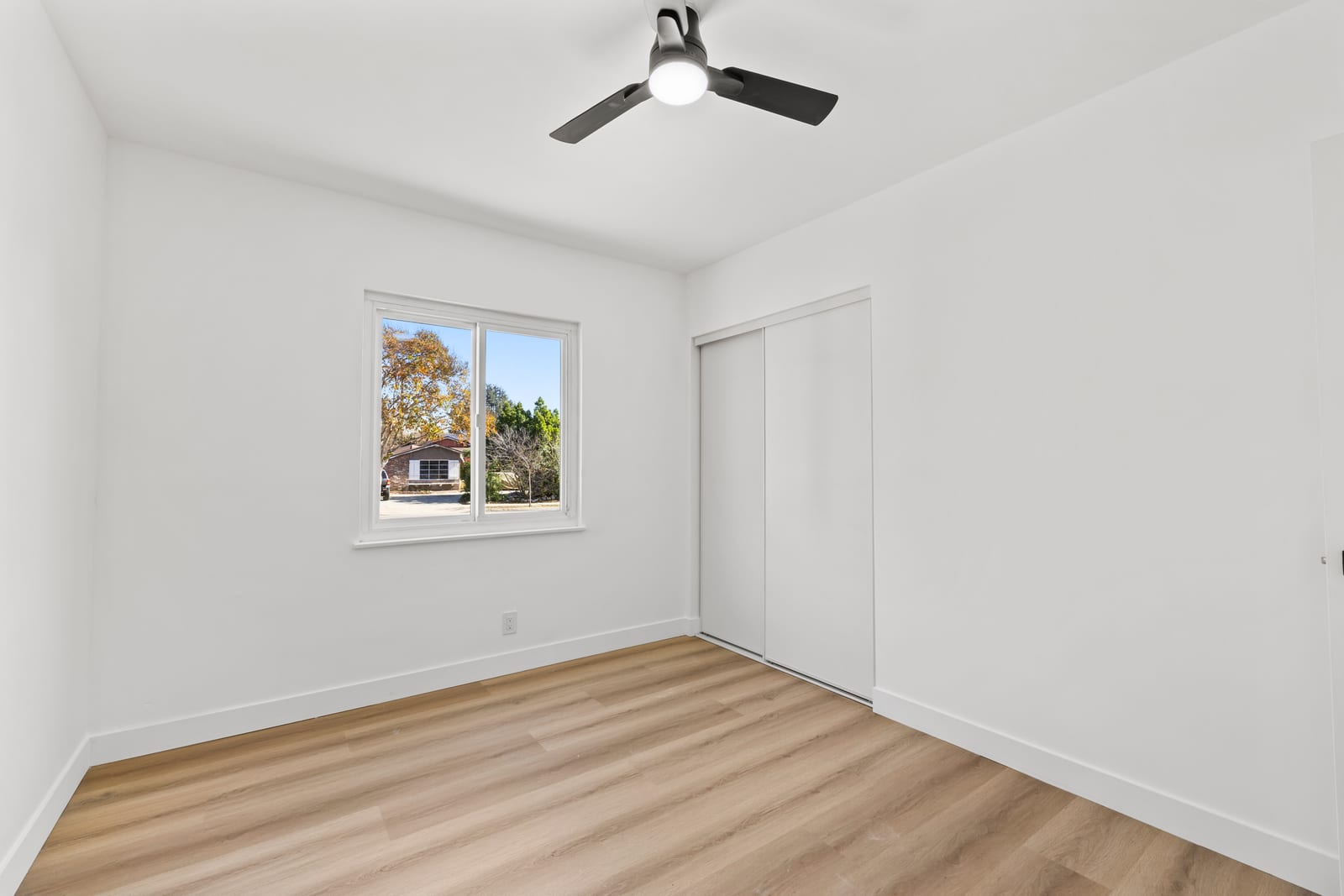 Empty bedroom in a Claremont home with light wood-look flooring, a white sliding closet door, a ceiling fan, and a window looking out on trees and a single-story house across the street