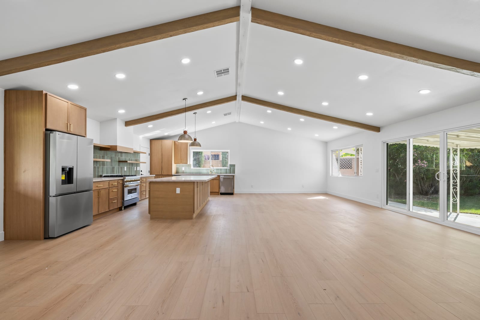 An empty open-plan great room with a vaulted ceiling and exposed wood beams, a wood-cabinet kitchen with an island and stainless appliances at the left, light plank flooring and sliding glass doors to a covered patio and garden at the right.