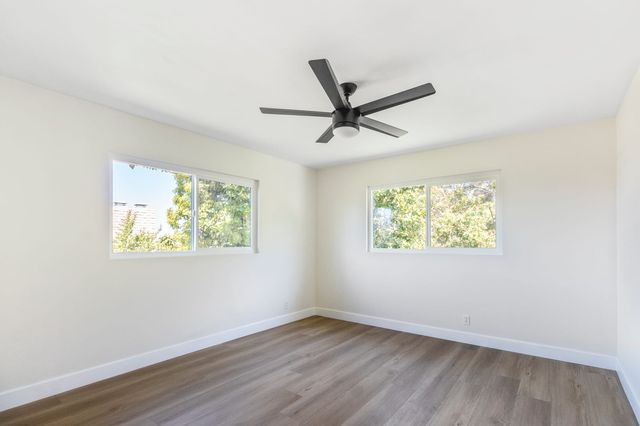 An empty corner bedroom with off-white walls, white baseboards, medium-brown plank flooring, a five-blade ceiling fan and two sliding windows framing green trees outside.