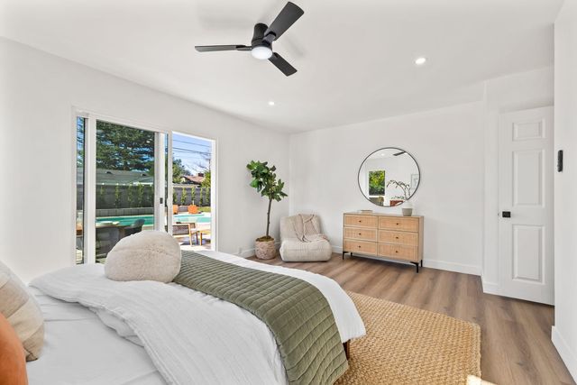 A bright bedroom with a ceiling fan, a made bed with a green quilt, a rattan-front dresser under a round mirror, and a sliding glass door open to a backyard pool.