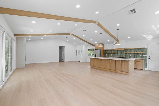 An empty open-plan great room with light wood-look flooring, exposed beams on a vaulted ceiling, and a wood kitchen island with a green tile backsplash beyond.