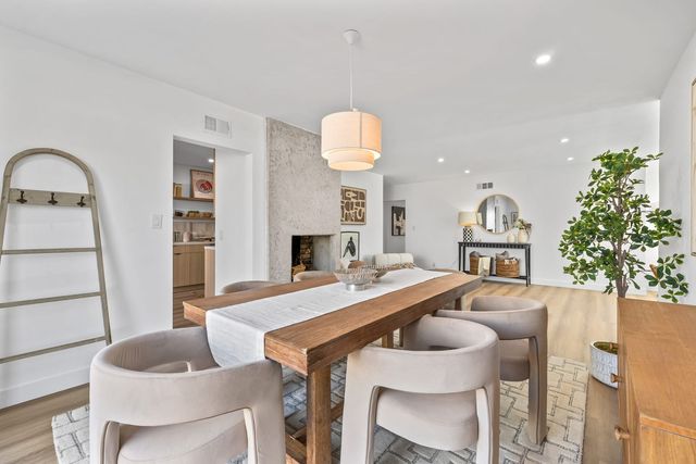 A dining room with a long wood table, upholstered chairs and a drum pendant light, with a plaster fireplace and a doorway to the kitchen behind.