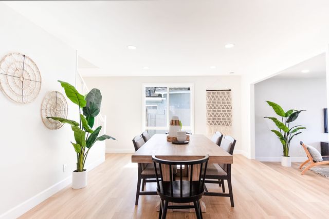A staged dining area with a long wood table, black spindle-back chairs, two tall potted plants and woven wall hangings in a bright room with light wood-look floors.