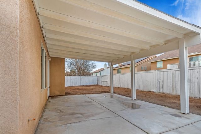 A covered back patio with a white wood patio cover over a concrete slab with rain puddles, a bare dirt yard and a weathered white fence, with neighboring two-story stucco houses beyond.