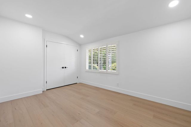 An empty bedroom in a Claremont home with light wood-look flooring, white walls, a sloped ceiling with recessed lights, white double closet doors and a shuttered window onto green trees.
