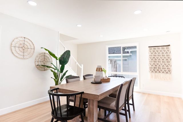 A staged dining area in a two-story house with a wood table and dark chairs, a white stair railing, woven wall hangings and a sliding window onto a side yard and the neighboring roofline.