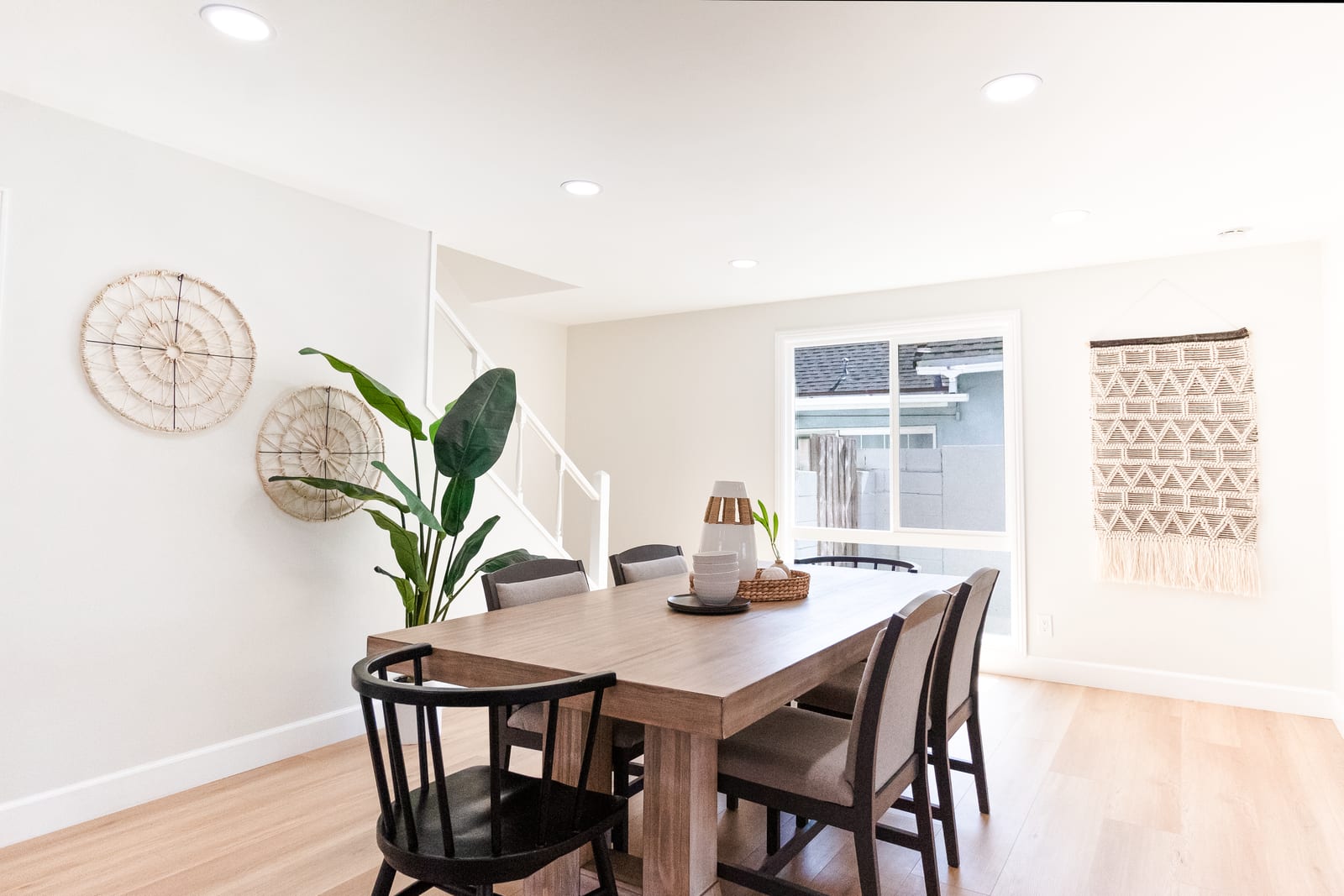 A staged dining area in a two-story house with a wood table and dark chairs, a white stair railing, woven wall hangings and a sliding window onto a side yard and the neighboring roofline.