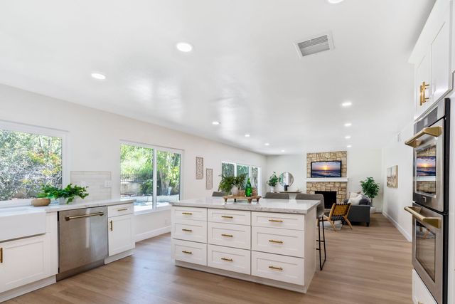 An open kitchen with a white island and brass pulls, double wall ovens and a farmhouse sink, leading to a family room with a stone fireplace, with windows onto a sloped, planted yard.