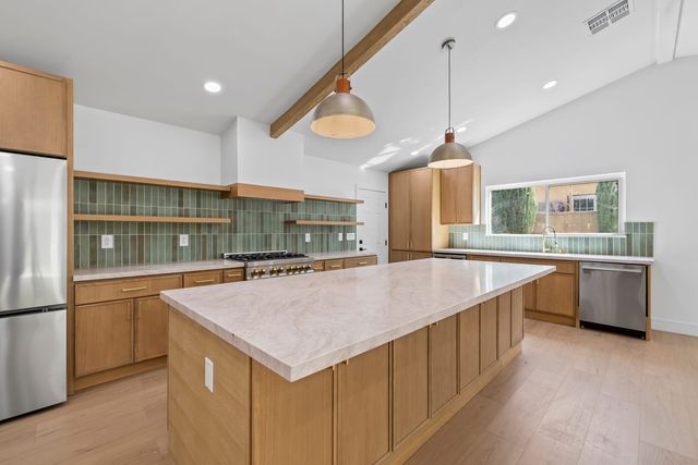 A remodeled kitchen in a Claremont house with oak cabinets, a long pale stone island, green vertical tile with open shelves, two hammered metal pendants and a wood beam across a vaulted ceiling