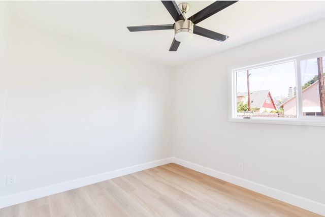 An empty bedroom with white walls, light wood-look flooring, a five-blade ceiling fan and a sliding window looking out at neighboring houses and utility poles