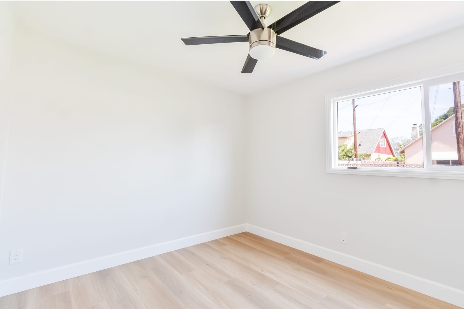 An empty bedroom with white walls, light wood-look flooring, a five-blade ceiling fan and a sliding window looking out at neighboring houses and utility poles