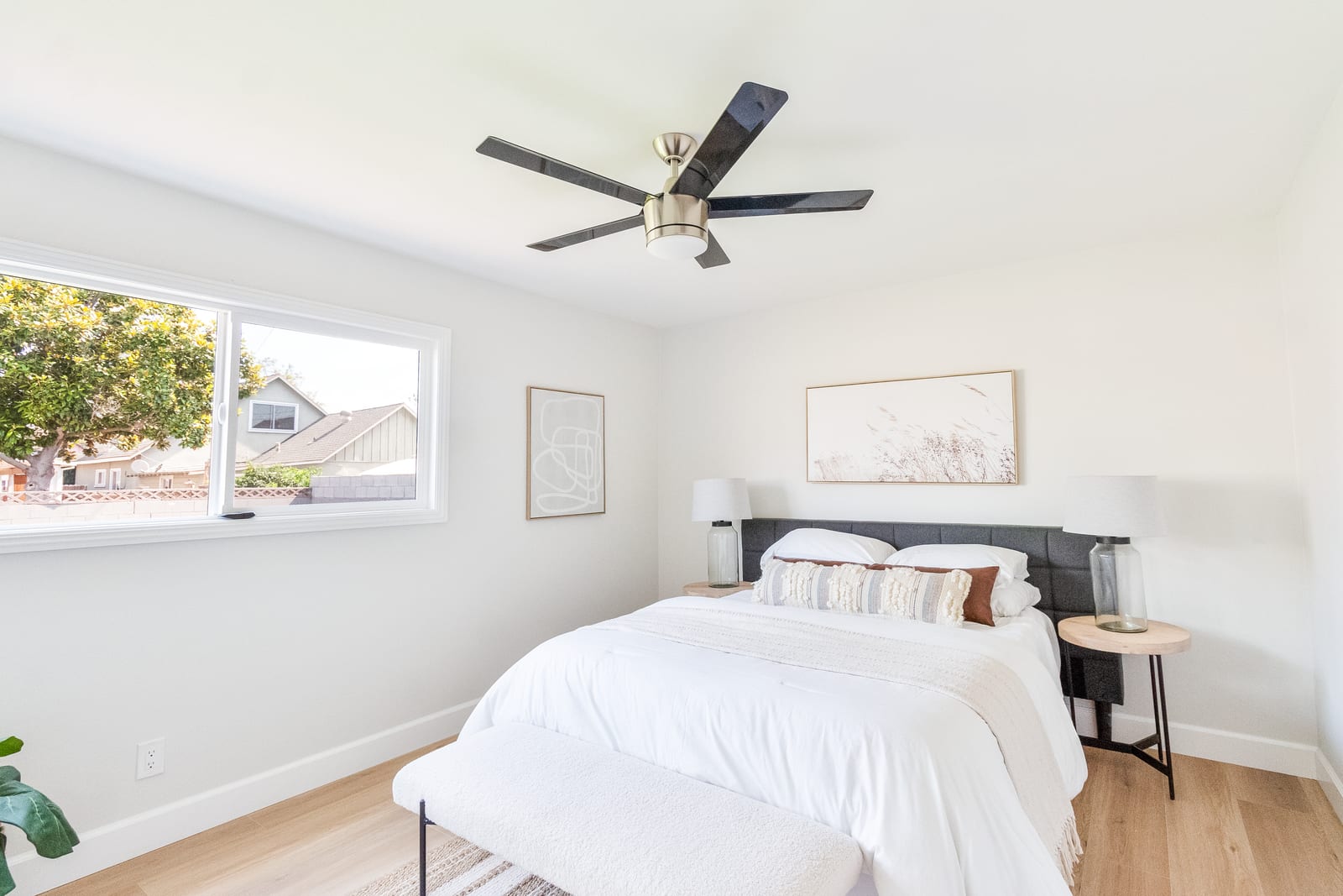 Bedroom with a ceiling fan, a made bed and a window looking out to neighboring rooftops and a tree