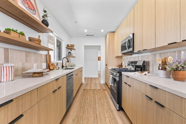 Galley kitchen in a Claremont home with light wood cabinets, open wood shelving, a gas range and a woven runner