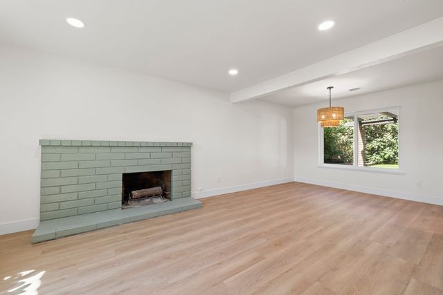 Empty living room with a painted green brick fireplace, light wood-look plank flooring, recessed ceiling lights and a woven pendant light by a window onto a garden.