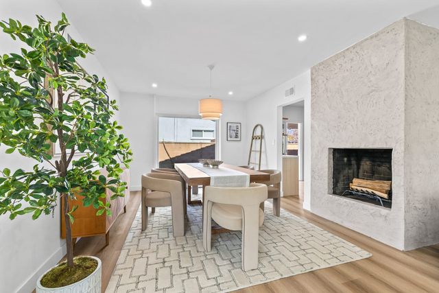 Staged dining room with a wood table and upholstered chairs on a patterned rug, a textured plaster fireplace holding cut firewood, and a potted tree beside a wood sideboard.