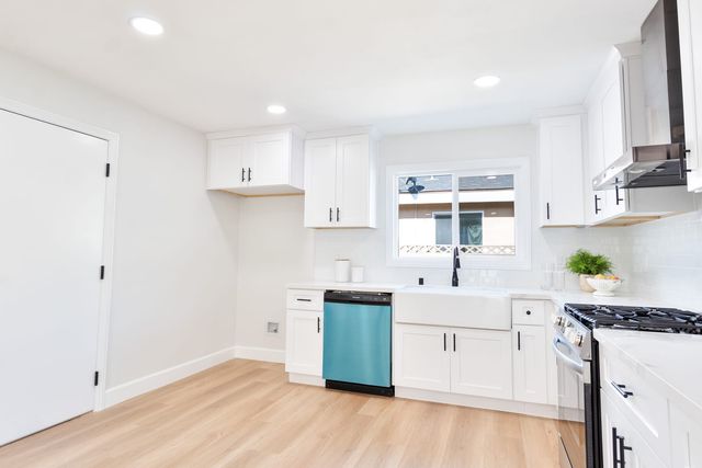 An empty, remodeled kitchen with white shaker cabinets, a farmhouse sink under a window, a gas range and light wood-look flooring.