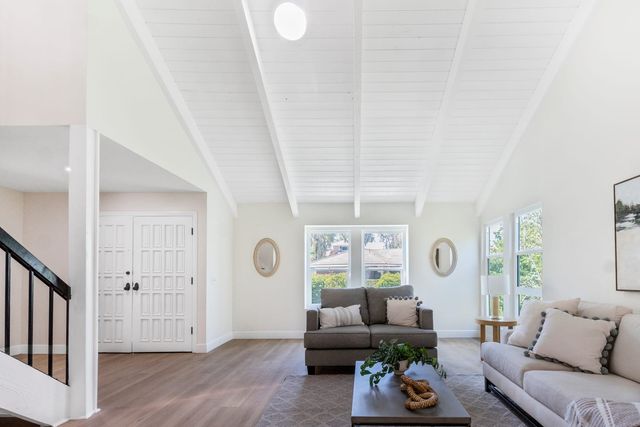 A staged living room with a vaulted white beamed ceiling, a skylight, double front doors and a staircase on the left.