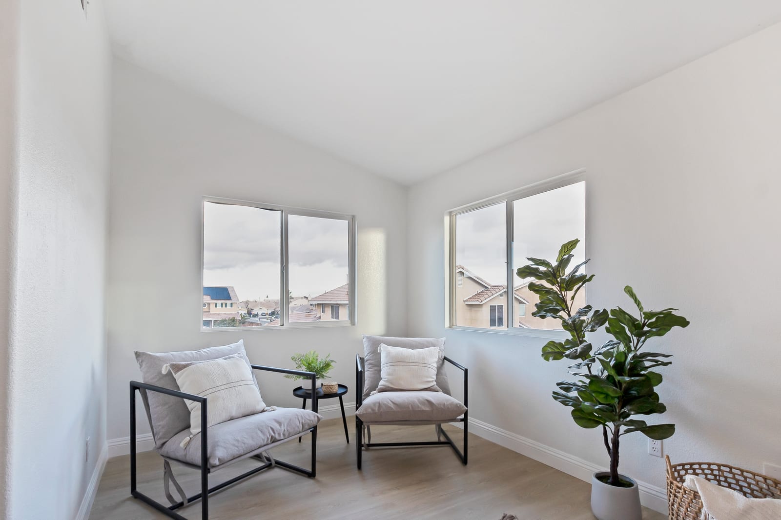 A small staged room with two armchairs and a potted plant, its windows looking out over neighboring two-story tract houses with tile roofs.
