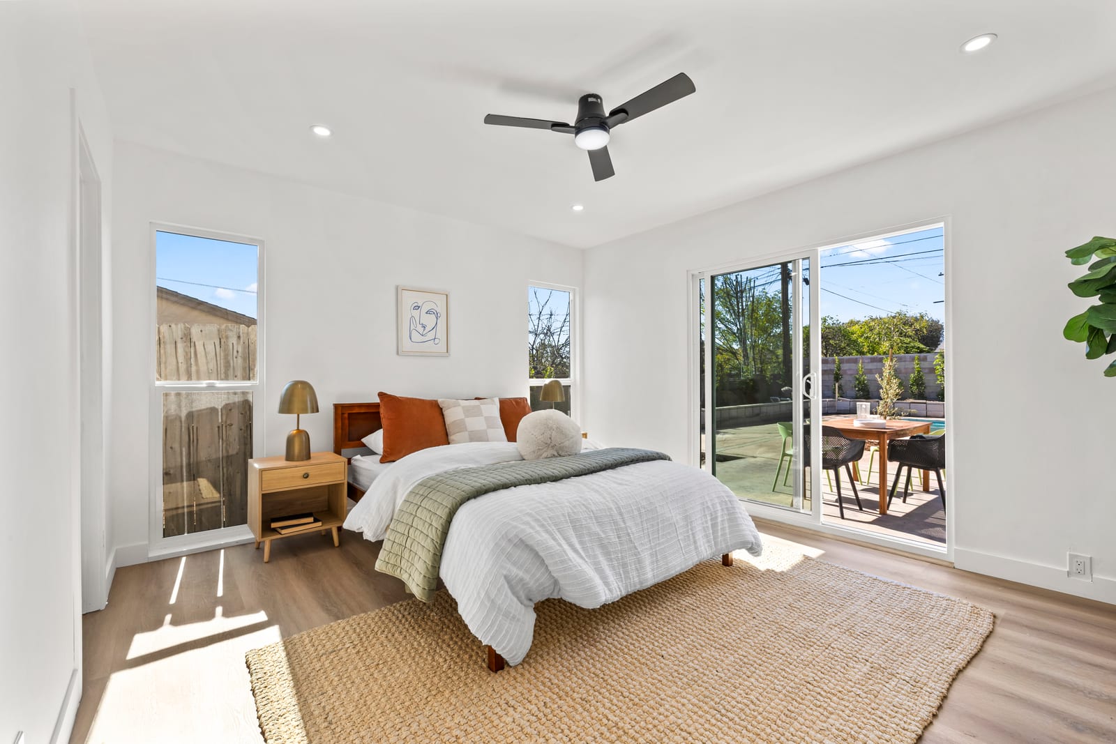 Bright bedroom with a made bed, a wood nightstand and a ceiling fan, sliding glass doors opening to a patio with a dining table