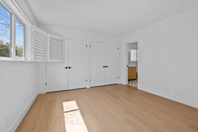 Empty bedroom with light wood floors, two sets of white closet doors, open plantation shutters and a doorway to a bathroom