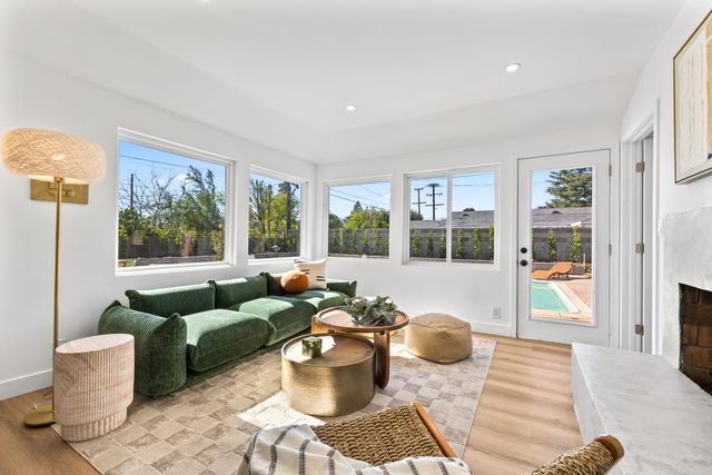 A staged living room in a Claremont house with a green velvet sofa, round wood and brass coffee tables, a white fireplace and corner windows facing a block-wall back yard, with a glass door opening to a small pool.