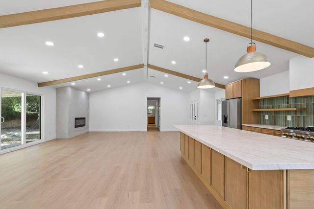 An empty, open great room in a Claremont single-story house under a vaulted ceiling with wood beams, a long stone-topped kitchen island with wood cabinets and green tile, and a sliding door to the yard.