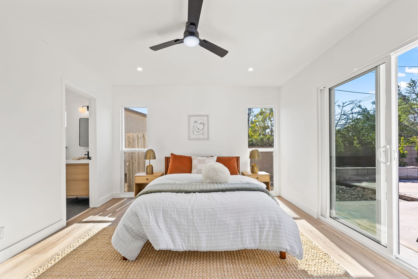 A staged bedroom in a Claremont house with a white-covered bed, brass bedside lamps, a black ceiling fan and a sliding glass door to a concrete patio and back yard.