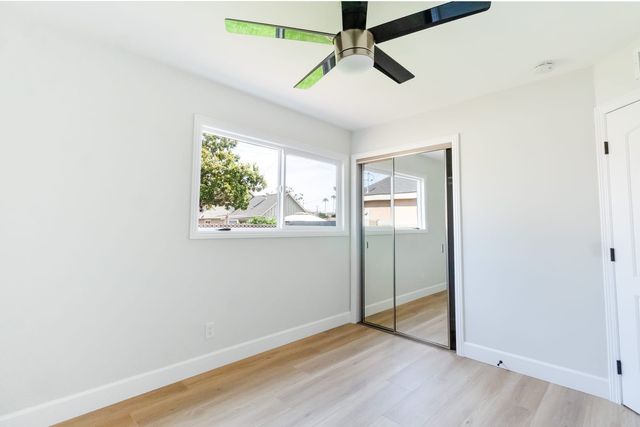 An empty bedroom with light wood-look flooring, a ceiling fan, a mirrored closet door and a window looking out at the pitched roofs of neighboring single-story houses.