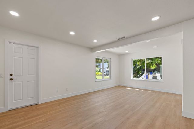 Empty living room with light wood floors, recessed lights, a white paneled door and two windows facing a green front yard