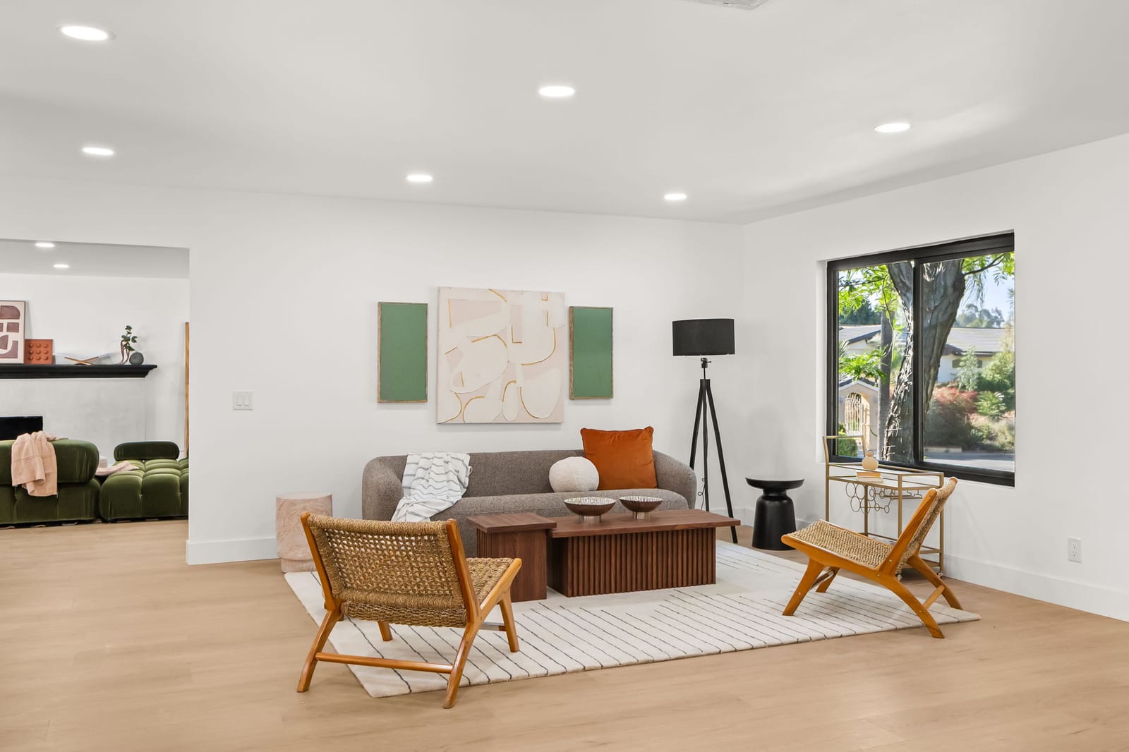 Staged living room with a gray sofa, two woven lounge chairs and a wood coffee table on a striped rug, a large window looking out to trees