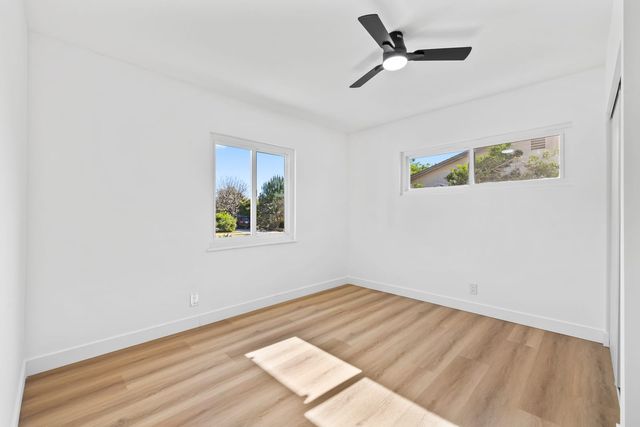 Empty bedroom with white walls, light wood-look plank flooring, a black ceiling fan and two windows looking out to trees and a neighboring roofline