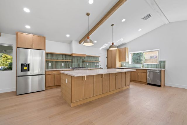 Empty remodeled kitchen with an oak-cabinet island, stainless refrigerator and dishwasher, green tile backsplash and two pendant lights under a beamed vaulted ceiling