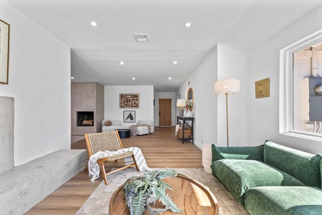 A long, bright living room with wide-plank wood floors, a green velvet sofa, a woven chair and a textured plaster fireplace, opening to a front entry door at the far end