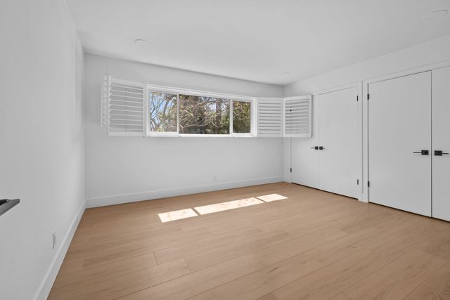 An empty bedroom with light oak plank floors, white walls, a wide window with open white plantation shutters framing a tree outside, and two pairs of white closet doors