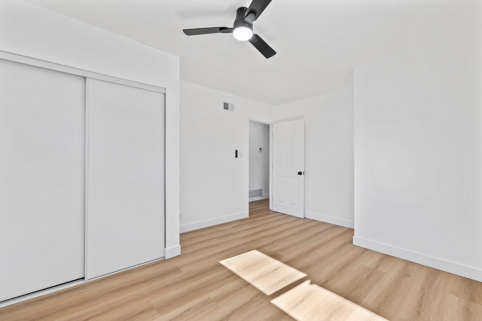 An empty bedroom with light wood-look plank floors, white walls, a white sliding closet, a black ceiling fan with a light, and an open six-panel door leading to a hallway