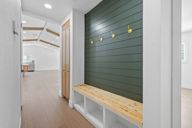 Built-in entry bench with open cubbies below a dark green shiplap wall with brass coat hooks, beside a hallway leading to an open kitchen